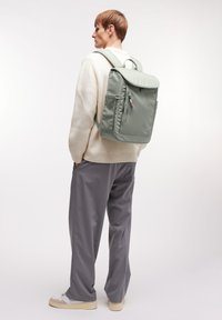 Hellgrauer Rucksack mit strukturierter Form, Reißverschlusstaschen und verstellbaren Trägern, aus strukturiertem Stoff gefertigt. Mit einem Logo auf der Vorderseite.