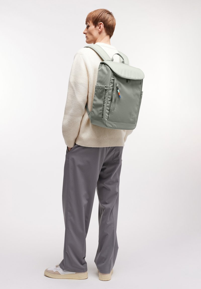 Personne portant un pull crème, un pantalon gris et des baskets blanches, avec un sac à dos gris clair doté d'une poche zippée à l'avant sur le dos.