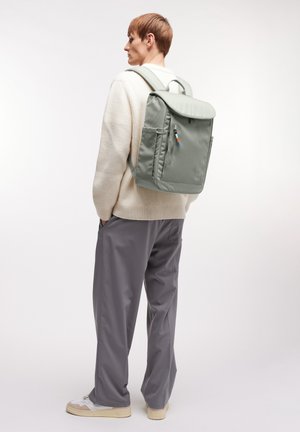 Persoon draagt een crèmekleurige trui, grijze broek en witte sneakers, met een lichtgrijze rugzak met voorvak met ritssluiting op de rug.