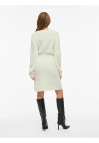 Kremasta pletena haljina sa dugim rukavima, strukirana remenom i crnim čizmama sa štiklom do koljena. Mekana tekstura, ravna silueta.