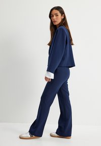 Costume bleu marine avec une veste courte et un pantalon évasé, présentant des détails de manchette blancs. Le modèle porte des baskets blanches, créant un look décontracté.