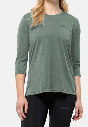 Kobieta ubrana w zieloną koszulkę z długim rękawem i czarne spodnie, obie z logo Jack Wolfskin, pokazana od szyi do połowy uda na białym tle.