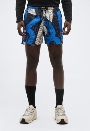 Homme portant un short à motifs bleu, noir et blanc, un haut à manches longues noir, des chaussettes noires et des chaussures de sport beiges, debout devant un fond blanc.