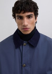 Junger Mann mit lockigem, dunklem Haar, der einen dunkelblauen Mantel mit einem Cordkragen und einem schwarzen, gerippten Rollkragenpullover vor einem einfarbigen Hintergrund trägt.