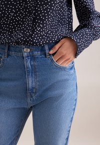 Jeans a vita alta in denim blu con una forma aderente, caratterizzati da una chiusura con bottone argentato e tasche laterali. Abbinati a una blusa blu navy con pois.