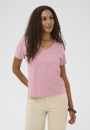 Vrouw met lang krullend haar die een roze T-shirt met V-hals en beige broek draagt, met één hand achter haar rug staand tegen een effen achtergrond.