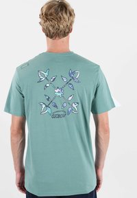 T-shirt en coton vert à manches courtes. Présente un motif floral en nuances de bleu et violet au dos. Marquage en dessous du motif.