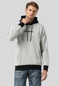 Hellgrauer Hoodie mit schwarzen Akzenten, Kordeln und Fronttasche. Mit Logo-Print auf der Brust. Weiches Material mit gerippten Bündchen.