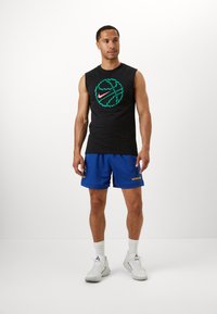 Fekete ujjatlan Nike póló zöld és rózsaszín kosárlabda grafikával, kék hálós sorttal, amelyen sárga "WARRIORS" felirat található. Fehér sportcipő.
