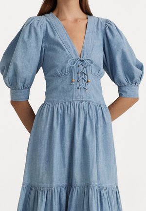 Frau in einem hellblauen Jeanskleid mit Puffärmeln, tiefem V-Ausschnitt und Schnürung vorne, von den Schultern bis zu den Knien gezeigt.