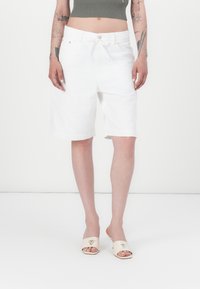 Hvite denimshorts med avslappet passform, med knapp- og snoråpning og oppbrettede kanter. Brukt med kremfargede sandaler.