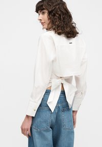 Femme aux cheveux bruns bouclés portant un chemisier blanc à manches longues noué avec un grand nœud dans le dos, associé à un jean bleu taille haute.