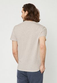 Uomo con capelli castani alla spalle, che indossa una camicia a maniche corte beige maculata e pantaloni blu, di spalle con una mano in tasca.