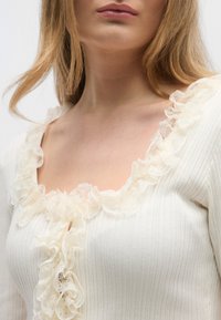Λευκό ριμπ τοπ με χαμηλή λαιμόκοψη και διακοσμητικό βολάν από δαντέλα, απαλής υφής και εφαρμοστή σιλουέτα.