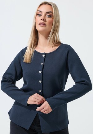 Blondynka w granatowym swetrze na guziki zapinająca dolny guzik na jednolitym jasnym tle.