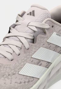 Gros plan sur une chaussure de course Adidas gris clair avec des lacets blancs et trois bandes blanches sur le côté, montrant la texture et les détails du logo.