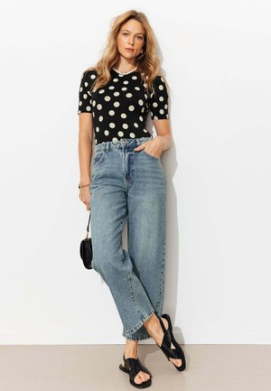 Femme blonde debout contre un mur blanc, portant un haut noir à pois, un jean bleu taille haute, des sandales noires, et tenant un petit sac noir.