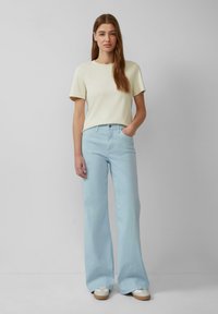 Helle gelbe Baumwoll-T-Shirt mit kurzen Ärmeln, kombiniert mit hochtaillierten, hellblauen, weit geschnittenen Jeans. Das Model steht mit einer Hand in der Tasche.