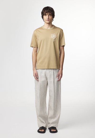 Beige bomullströja med ett blommotiv på bröstet, kombinerad med lösa randiga byxor i kräm och svart, samt svarta slide-sandaler.