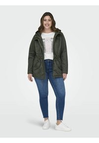 Olivgrüne Parka-Jacke mit Kordelzug in der Taille, Reißverschluss-Taschen und Kapuze, getragen über einem weißen T-Shirt mit Grafik und blauen Jeans. Weiße Sneaker.