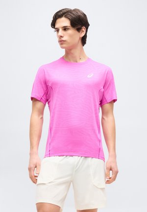 Fiatal férfi élénk rózsaszín rövid ujjú sportpólóban és fehér rövidnadrágban, semleges világos háttér előtt áll.