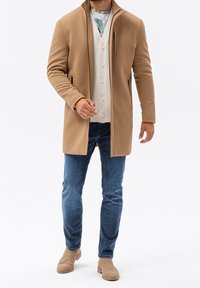 Tannfärgad ytterjacka med hög krage, sidofickor med dragkedja, lager på en krämfärgad skjorta med knappar och blå jeans, kompletterat med tannfärgade skor.