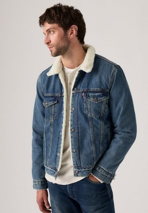 Chaqueta de mezclilla con un cuello forrado de piel sintética de color beige claro, diseño clásico de cinco bolsillos y un tono azul oscuro. Cuenta con botones metálicos y detalles de costura.