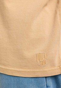 T-shirt en coton jaune à manches courtes avec logo brodé en bas. Texture lisse et design décontracté, associé à un jean en denim bleu clair.