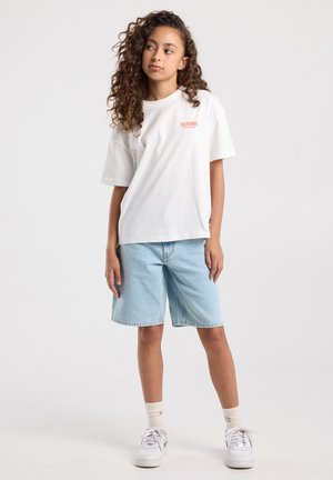 Tienermeisje met krullend haar, gekleed in een wit "California" T-shirt, lichte denim short, witte sokken en witte sneakers, staand tegen een effen achtergrond.