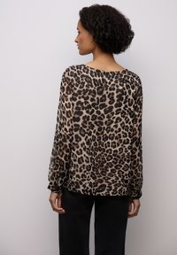 Przezroczysta bluzka z nadrukiem w cętki, zaokrąglony dekolt, długie rękawy z elastycznymi mankietami oraz luźny krój. Paleta kolorów obejmuje beż i czerń.