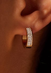 Gouden hoopsierra met een rij heldere, fonkelende edelstenen aan de voorkant. Gladde afwerking met een glanzende, metalen textuur.