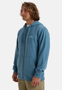 Haut à zip bleu en tissu doux, doté d'une poche frontale, d'une capuche avec cordon de serrage et d'un logo brodé sur la poitrine.
