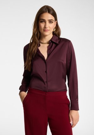 Donna con lunghi capelli castani indossa una camicetta abbottonata di colore viola scuro e pantaloni rossi a vita alta, in piedi con una mano in tasca su sfondo uniforme.