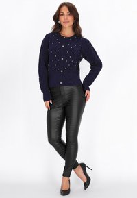 Laivansininen neulecardigan, jossa on helmikirjailuja, pyöreä kaula-aukko ja nappisulku, yhdistettynä kimaltaviin mustiin nahkahousuihin ja kärkitossuihin mustilla koroilla.