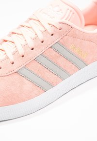 Růžové semišové tenisky se třemi šedými pruhy, texturovanými detaily a bílou gumovou podrážkou. Obsahují zlatý nápis "GAZELLE" poblíž  šněrování.