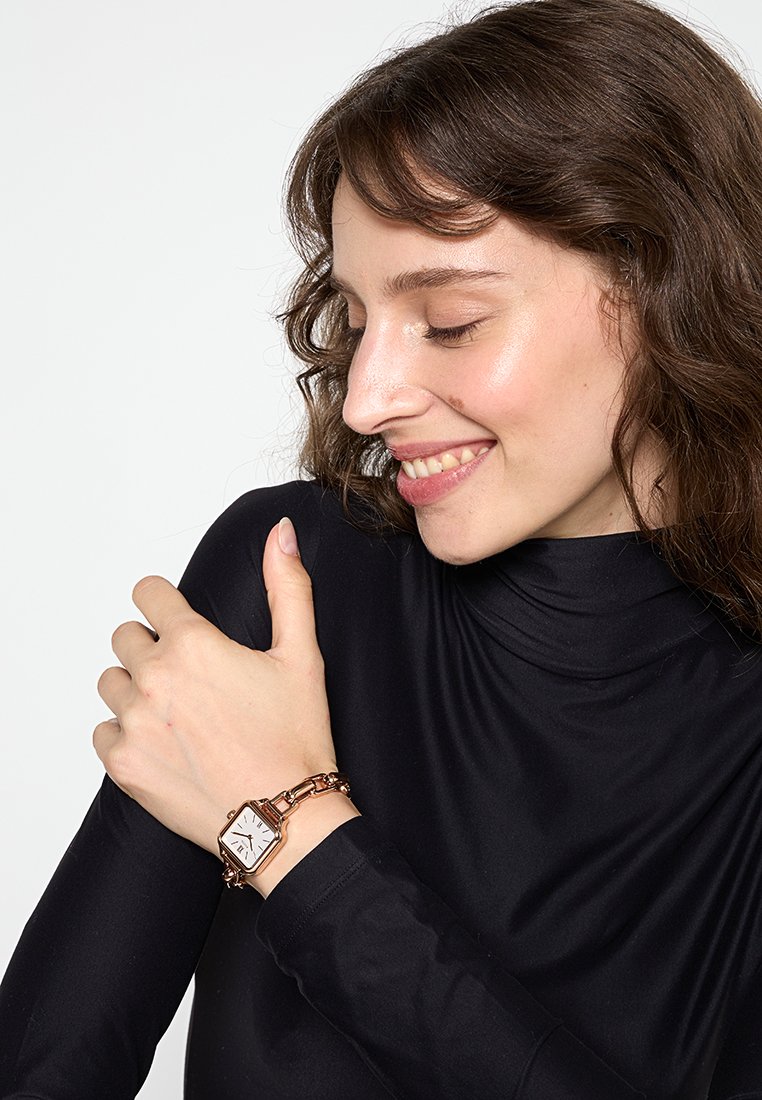 Orologio quadrato con quadrante bianco e dettagli in oro rosa. Collegato a un bracciale in oro rosa a catena. Indossato su una maglietta nera a maniche lunghe.