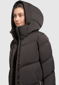 Black puffy jacket with a high collar and large hood. Features zip and snap closures, quilted padding, and side pockets for added functionality.