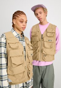 Beige Utility-Westen mit mehreren Taschen, aus leichtem Stoff gefertigt, kombiniert mit einem karierten Hemd und pastellfarbenen Schichten darunter.