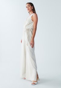 Lysebeige heldragt med dyb V-hals, snøre i taljen og vide, flagrende ben. Glat tekstur og sideslidser for ekstra detaljer.