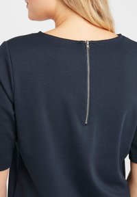 Tumedat sinise värvusega lühikeste varrukatega pluus, millel on seljaosal tõmblukk ja sile, tekstuuriga kangas. Lihtne, moodne disain.