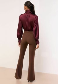Bluza de satin burgund cu guler înalt și mâneci bufante, asortată cu pantaloni evazați maro, cu talie înaltă. Texturi netede, design ajustat.