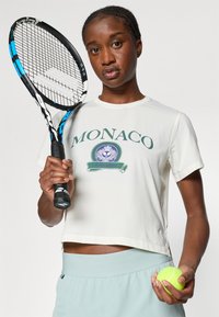 Fehér cropped top zöld "MONACO" felirattal és logóval. Világoskék sportnadrág; teniszező ütőt tart a kezében, valamint egy neonzöld teniszlabdát.