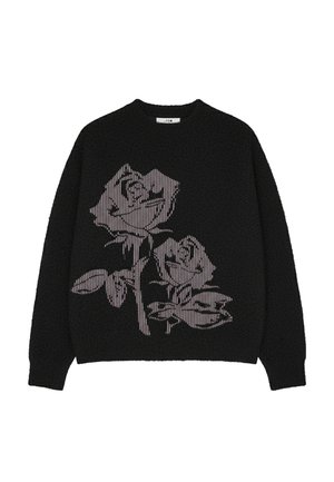 Young man wearing a black fuzzy sweater with a large purple rose design and white pants, standing against a plain gray background.