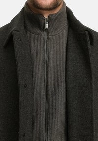 Grauer Wollmantel über dunkelgrauem, geripptem Reißverschluss-Pullover. Auffällige Merkmale sind der hohe Kragen und kontrastierende Texturen, sowie Knopfakzente am Mantel.