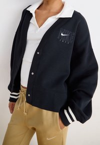 Marinblå sweatshirtsjacka med vit logotyp, rund hals, knappar och randiga muddar. Matchas med beige sweatpants med en logotyp.