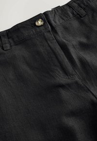 Gros plan de la ceinture d’un pantalon en lin noir avec un seul bouton foncé, des passants de ceinture et une couture au niveau de la poche latérale.