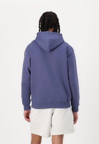 Hooded sweatshirt i enfärgad lila, tillverkad av mjuk, texturerad tyg. Har en känguruficka och ribbad muddar och fåll. Bärs med grå shorts.