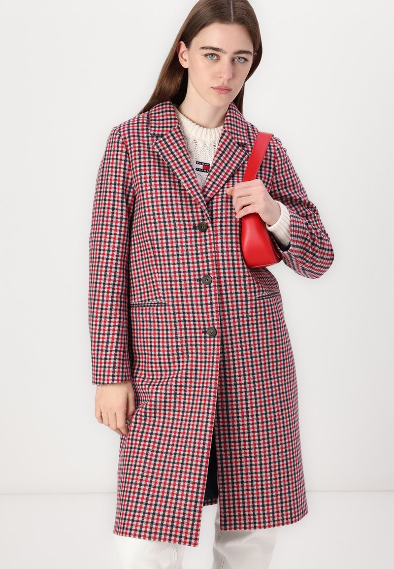 Czerwono-granatowy płaszcz w kratę z klapami, przednimi kieszeniami i trzema guzikami. Zestawiony z kremowym swetrem dzianinowym i czerwoną torebką.