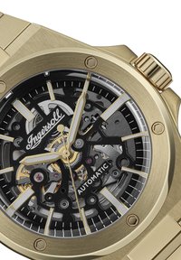 Goudkleurig automatisch horloge met een skeletdial met zwarte accenten, zilveren uur markers en een gestructureerde metalen armband.