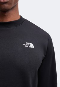 Gros plan d'une personne portant un sweat-shirt noir à col rond avec un logo blanc The North Face sur la poitrine gauche.